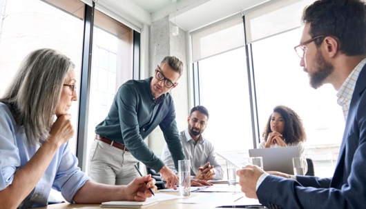 Business people around a conference table with bright windows in the background discussing documents.