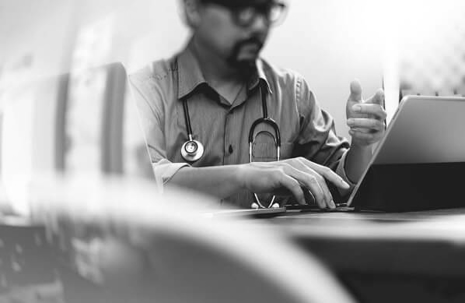 Black and white photograph of a physician in the background typing on a laptop. Objects in the foreground are blurred.