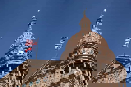 Texas capitol dome with United States and Texas flags blowing in breeze against blue sky.