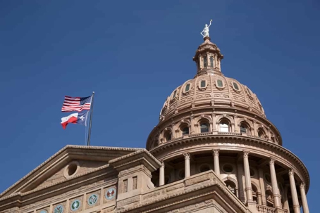 Texas capitol dome with United States and Texas flags blowing in breeze against blue sky.