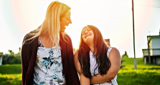 A woman walks outdoors with a female teenager with Down syndrome, smiling and laughing together.