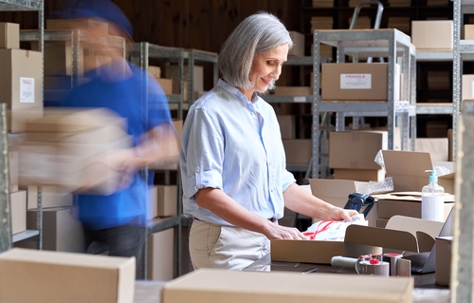Female small business owner packing an order in box in warehouse with her employee in blur motion behind her carrying boxes.
