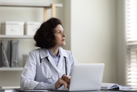 Stressed female physician looking in the distance, considering the consequemces of a False Claims Act investigation, sitting in front of a laptop in clinic office.