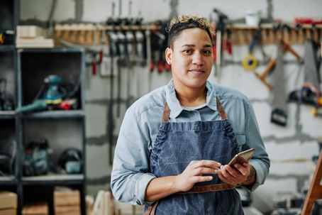 Manufacturing shop owner using her cell phone and looking confidently at camera.