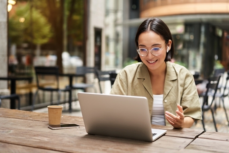 Young woman sitting on online meeting in outdoor cafe, talking to laptop camera.