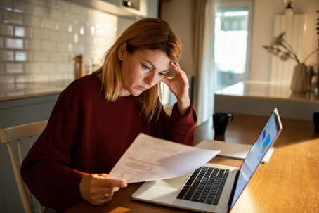 Concerned business owner holding financial statement and comparing it to figures on her laptop.