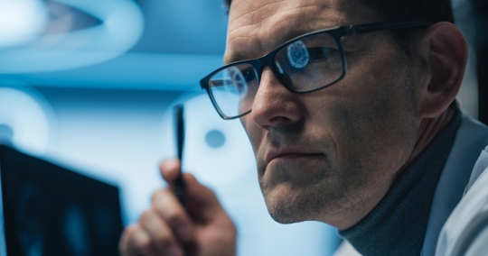 Physician examines AI-enhanced brain scans on computer screen reflected in his glasses.