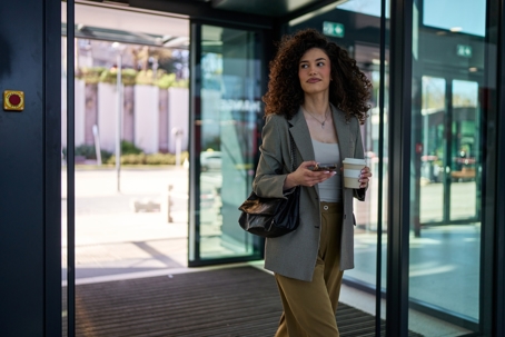 Businesswoman exiting an office building, holding a smartphone in one hand, looking away with confidence.