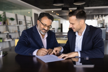 Two businessmen in suits sitting at table in office signing a letter of intent (LOI).