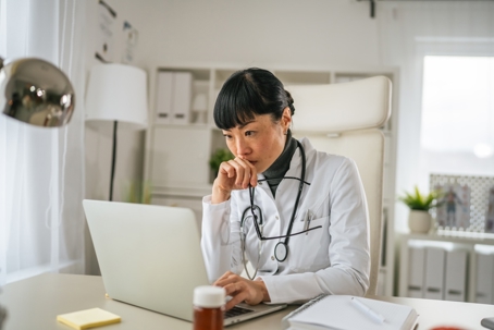 Doctor wearing a white lab coat and stethoscope, analyzing medical billing data on a laptop in her medical office.