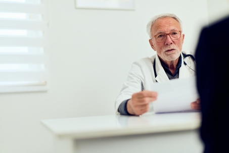 Elderly male doctor in a hospital office reviews documents across a table from a peer.