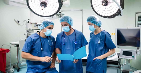 A group of physicians in surgical scrubs discuss a patient in an operating room..