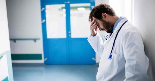 A doctor in a white lab coat leans against a hospital wall, holding his forehead with one hand, looking upset.