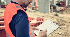 An OSHA compliance safety and health officer wearing an orange safety vest inspects a construction site completing a form clipped to a clipboard.