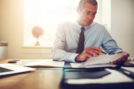 An executive reviews a printed purchase agreement with a pen in hand. A sunny window is in the background.