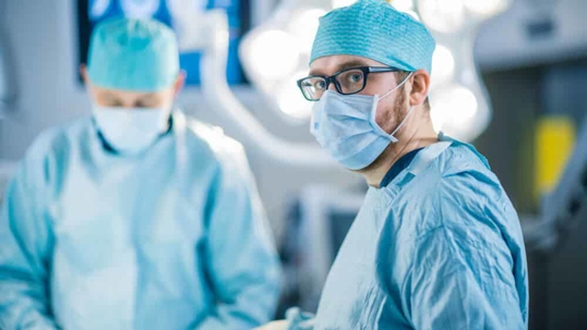 A surgeon wearing glasses and scrubs looks into the camera with a fellow surgeon and operating room in the background.