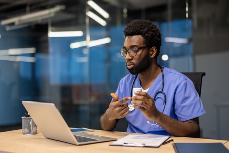 An African American doctor in scrubs explaining the medication uses during a telehealth appointment in his office, using a laptop.