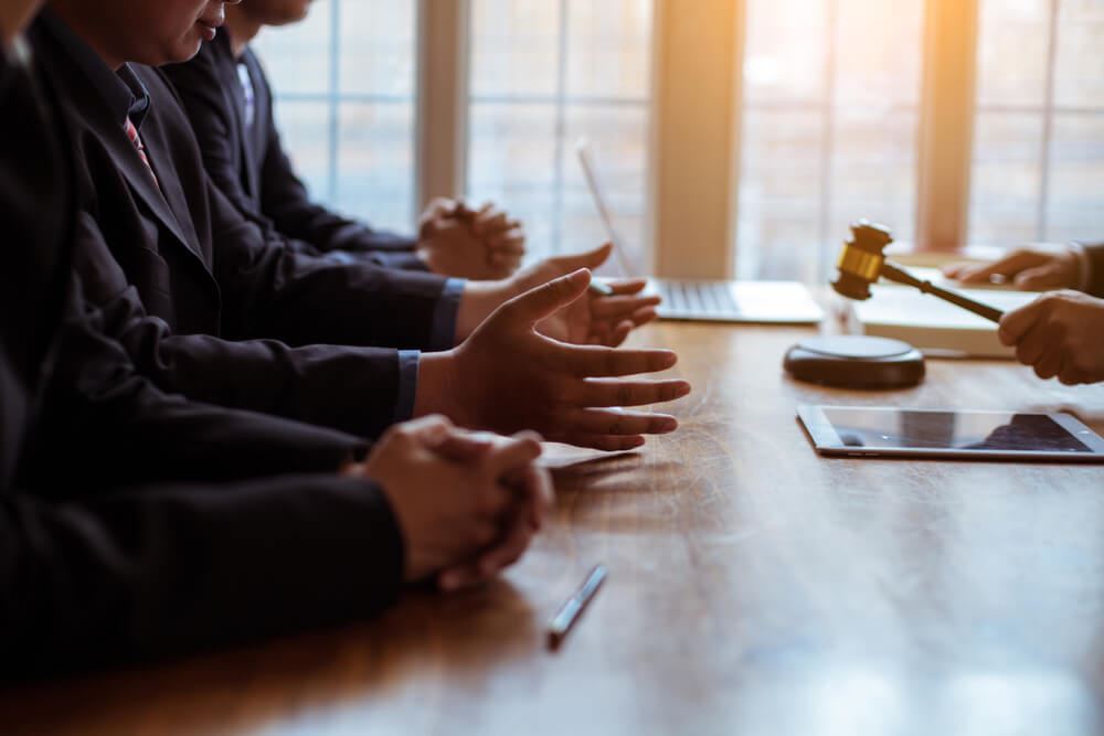 Businessmen seated at a conference table present their case before an arbitrator holding a gavel.